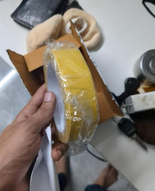 Hand holding a yellow roll of heavy duty carpet adhesive tape wrapped in plastic over a cardboard box