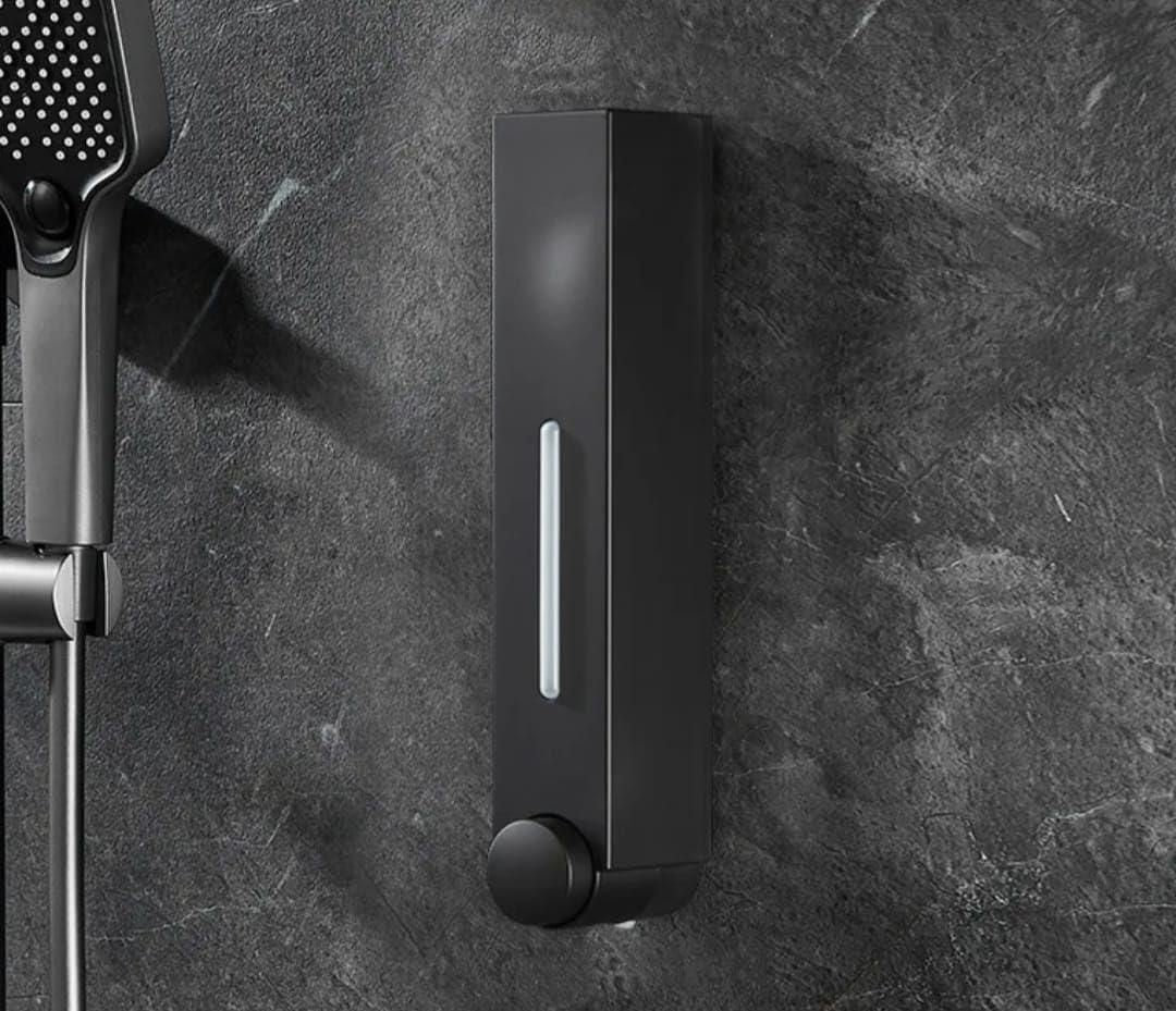 Matte black wall mounted soap dispenser with clear level window and round pump knob next to handheld shower head