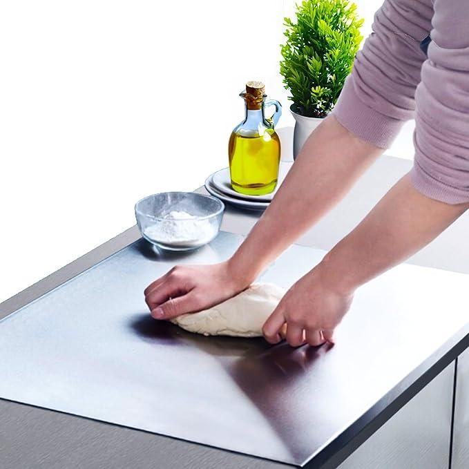 Person kneading dough on a stainless steel chopping board in a kitchen setup with olive oil and a plant