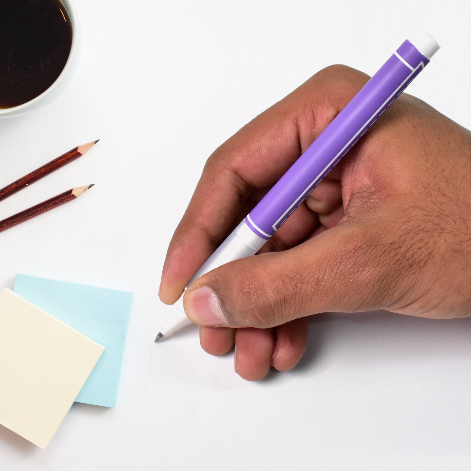 Hand holding a Your Brand 0.5 mm black ball pen over sticky notes and a coffee cup