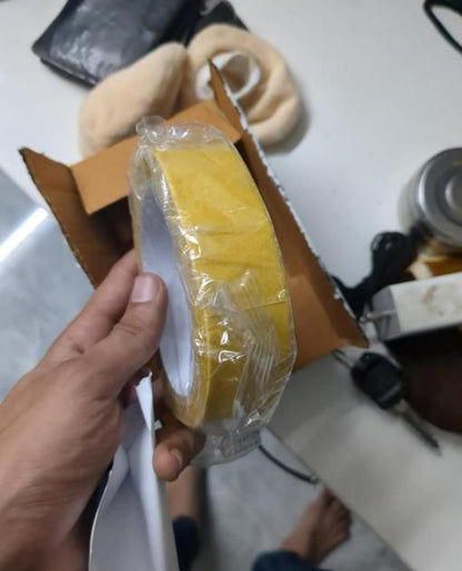 Hand holding a yellow roll of heavy duty carpet adhesive tape wrapped in plastic over a cardboard box