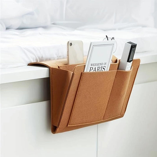 Brown bedside pocket organizer for essentials hanging over bed frame with phone, glasses, and remote