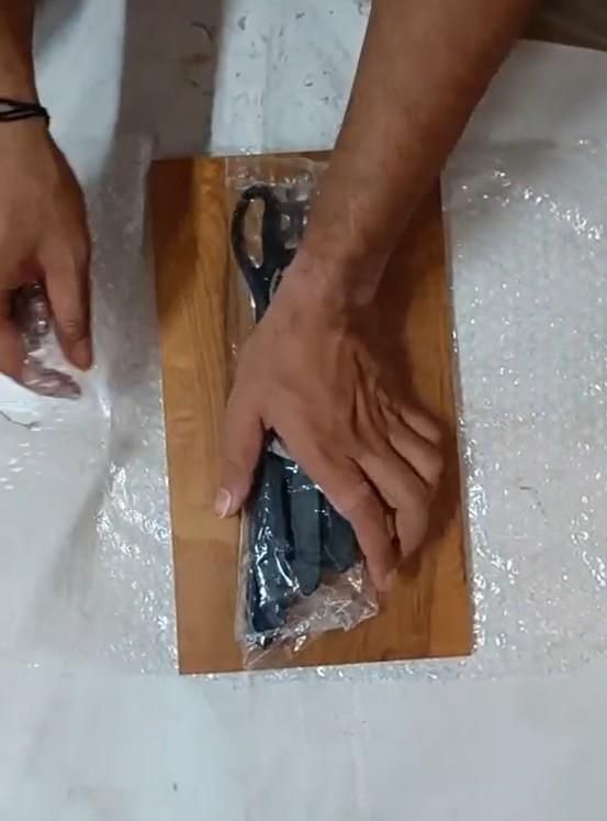 Top view of hands unwrapping wooden chopping board with knife set in plastic packaging on table