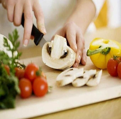 Person slicing mushroom on a wooden chopping board with knife set surrounded by cherry tomatoes yellow bell pepper and herbs