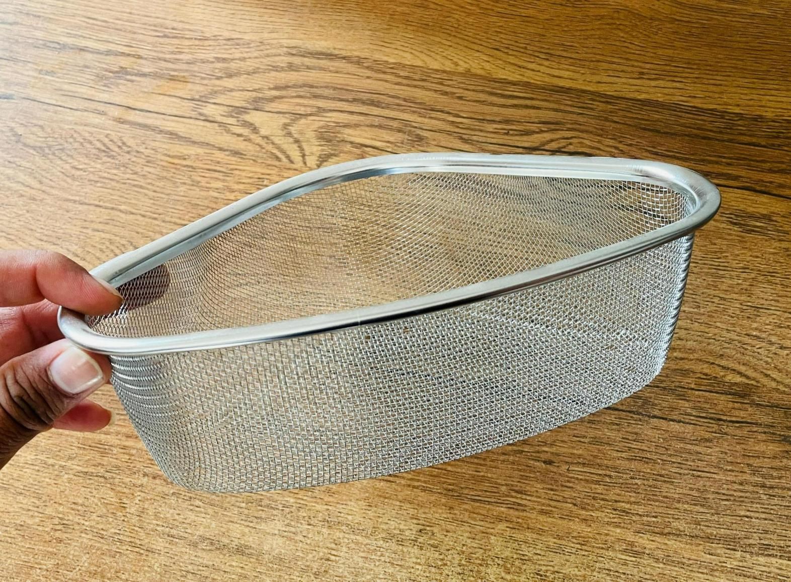 Stainless steel corner sink strainer for kitchen held over wooden surface by hand