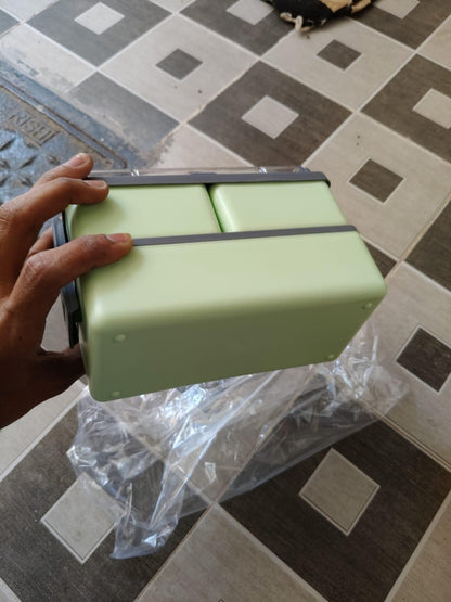 Hand holding a green bento compartment lunch box with multiple compartments on tiled floor