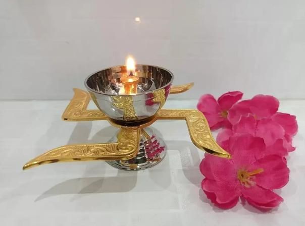 Handcrafted puja brass diya with lit flame and decorative gold-toned details next to pink flowers